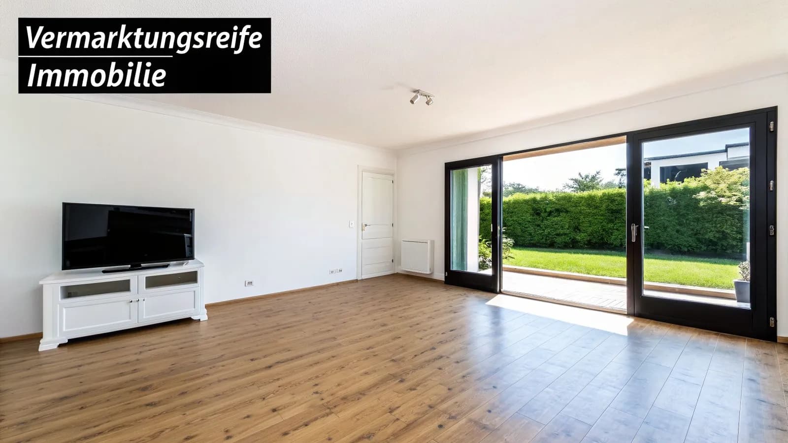 Helles, leeres Wohnzimmer mit Holzboden, weißem TV-Schrank und Schiebetüren zum grünen Garten.