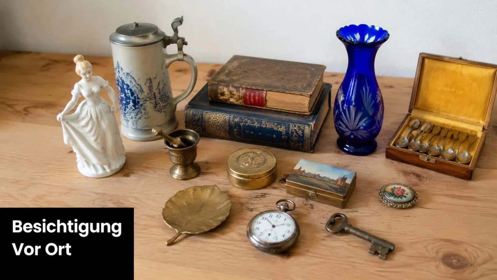 Verschiedene antike Gegenstände wie Bücher, Porzellan, Besteck und eine Taschenuhr liegen dekorativ auf einem Holztisch.