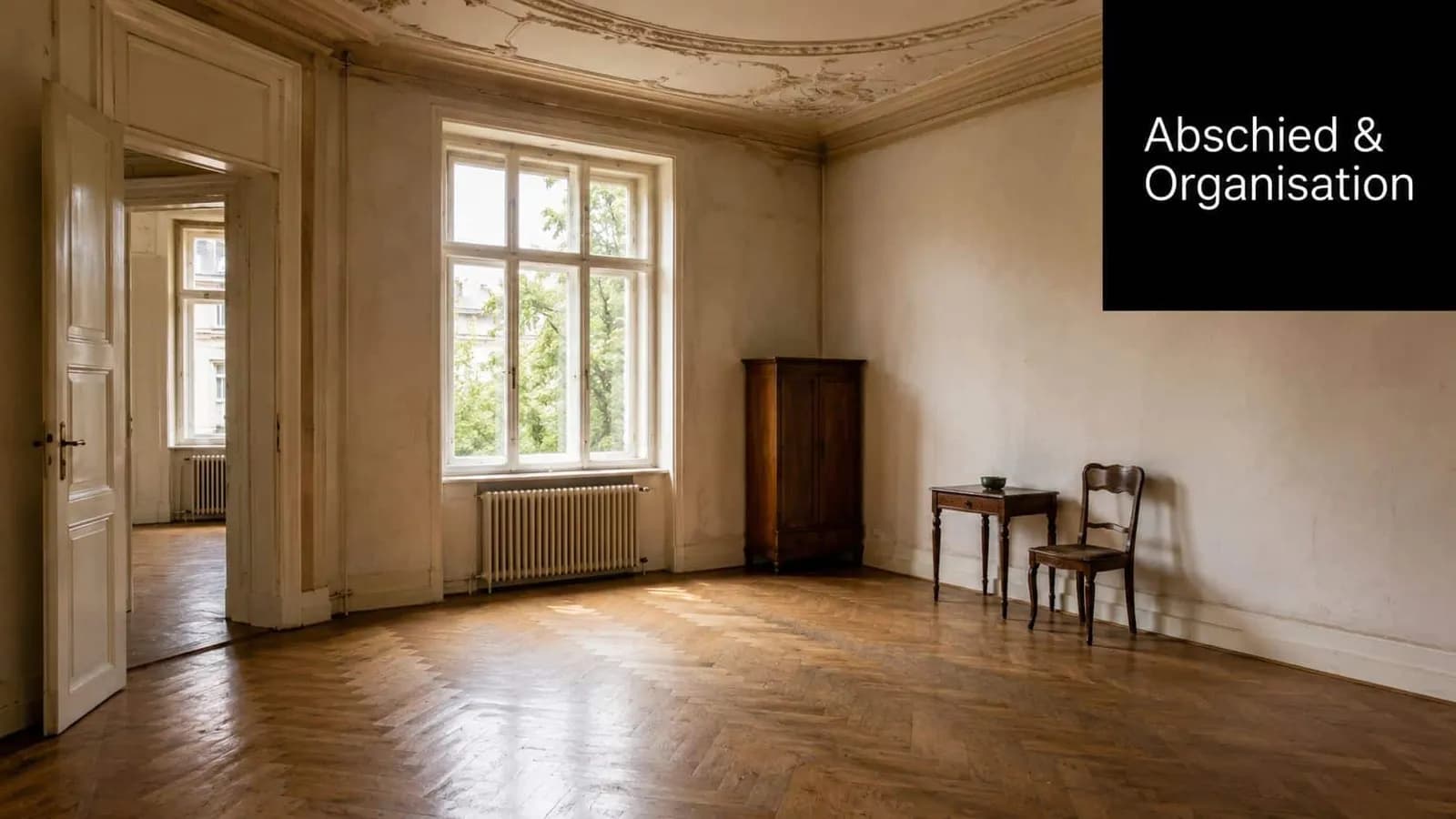Ein geräumiges, leeres Zimmer mit Fischgrätparkett, einem Fenster, einem alten Holzschrank sowie Tisch und Stuhl.