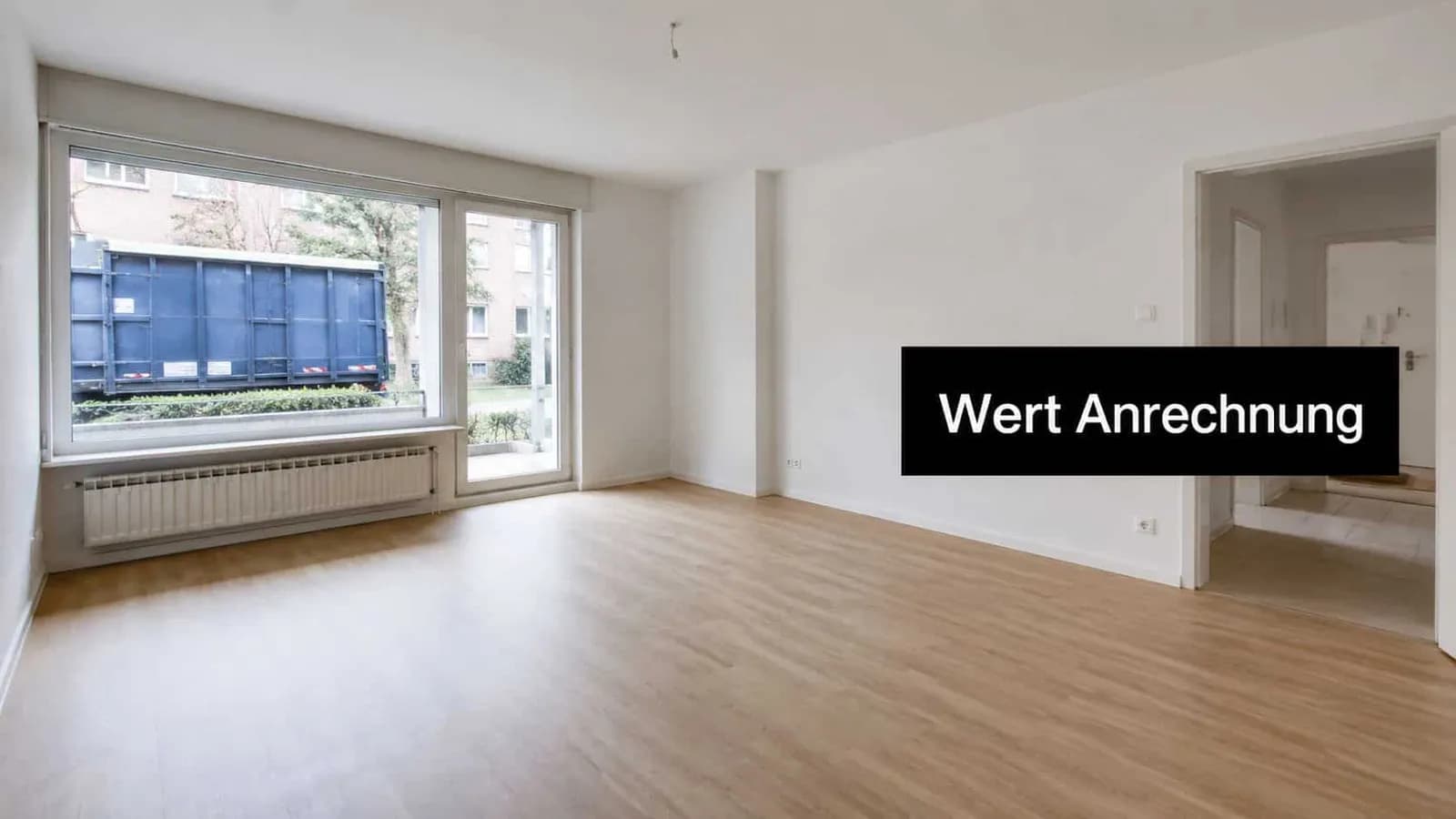 Ein helles, leeres Zimmer mit weiß gestrichenen Wänden, Holzboden, großem Fenster und offener Tür zur Wohnung.